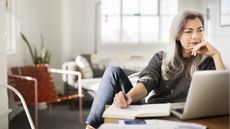A woman looks thoughtful as she reads something on her laptop and takes notes.